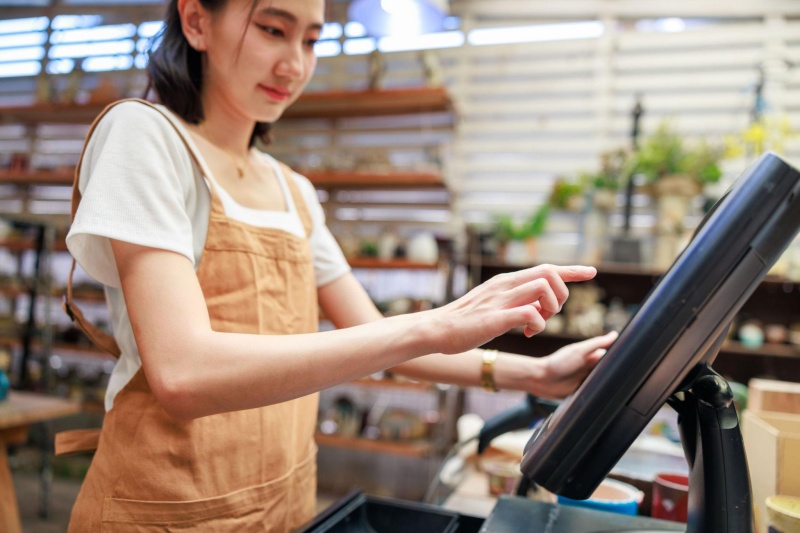 Retail Staff Using a POS System for Sales Transactions ภาพพนักงานร้านค้ากำลังใช้งานเครื่อง POS แบบหน้าจอสัมผัสเพื่อจัดการรายการขายภายในร้าน แสดงถึงการนำเทคโนโลยีระบบขายหน้าร้านมาใช้ในการบันทึกข้อมูลสินค้าและอำนวยความสะดวกในการทำธุรกรรม เหมาะสำหรับประกอบบทความเกี่ยวกับระบบ POS การจัดการงานขาย และข้อควรรู้เกี่ยวกับ ภ.พ.06 สำหรับผู้ประกอบการร้านค้า
